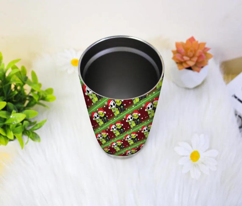 May include: A stainless steel tumbler with a wraparound pattern of green creatures wearing Santa hats on a red and green striped background. The tumbler has a black interior and a silver rim. The design is repeated diagonally.