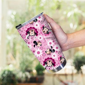 以下が含まれることがあります： ミニーマウスのキャラクターと白いデイジーの花が繰り返されるピンク色のタンブラー。タンブラーはステンレス製のベースと黒い蓋が付いています。「ミニー」の文字が筆記体で書かれています。