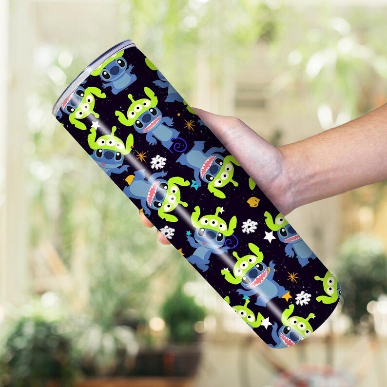Peut inclure: Un gobelet en acier inoxydable avec un motif r&eacute;p&eacute;t&eacute; de personnages de dessins anim&eacute;s extraterrestres bleus et Stitch sur fond noir. Le motif comprend des &eacute;toiles blanches et des accents jaunes. Le gobelet est tenu dans une main.
