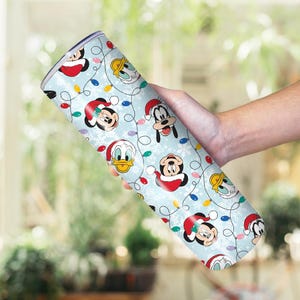 May include: A light blue tumbler featuring cartoon characters Mickey Mouse, Donald Duck, and Goofy wearing Santa hats. The tumbler is decorated with Christmas lights and snowflakes.
