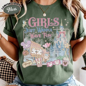 May include: Moss green t-shirt with the text "GIRLS JUST WANNA HAVE FUN" in pink and white. The graphic includes a castle, fireworks, and various Disney-themed icons. The shirt is a Comfort Colors brand.