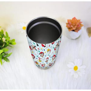 May include: A stainless steel tumbler with a light blue background and a repeating pattern of cartoon characters wearing Santa hats and Christmas lights. The tumbler is cylindrical and has a dark interior.