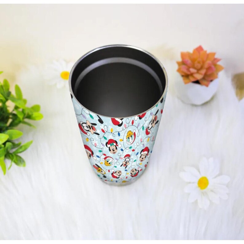 May include: A stainless steel tumbler with a light blue background and a repeating pattern of cartoon characters wearing Santa hats and Christmas lights. The tumbler is cylindrical and has a dark interior.