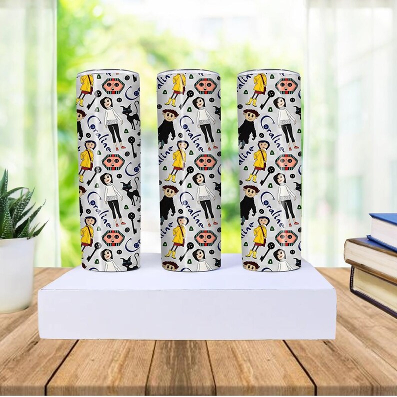 May include: Three stainless steel tumblers with a repeating pattern of a girl in a yellow raincoat, a black cat, and the word "Coraline" in a script font. The background is light gray.