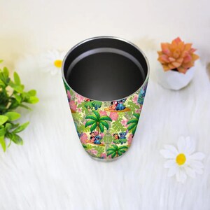 May include: A stainless steel tumbler with a colorful tropical design featuring cartoon characters, palm trees, and flowers. The tumbler has a black interior and a silver rim. The design includes shades of green, pink, and blue.