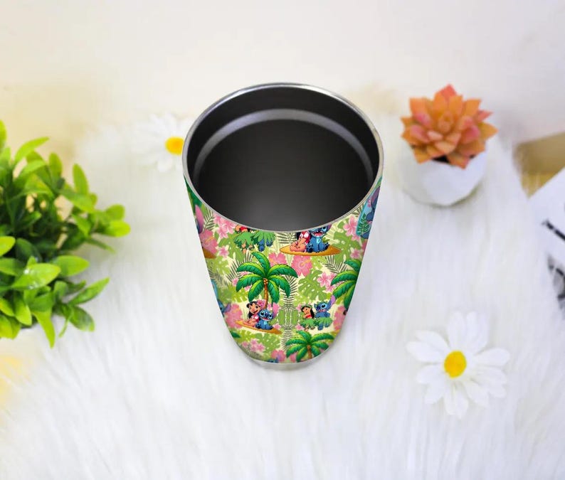 May include: A stainless steel tumbler with a colorful tropical design featuring cartoon characters, palm trees, and flowers. The tumbler has a black interior and a silver rim. The design includes shades of green, pink, and blue.