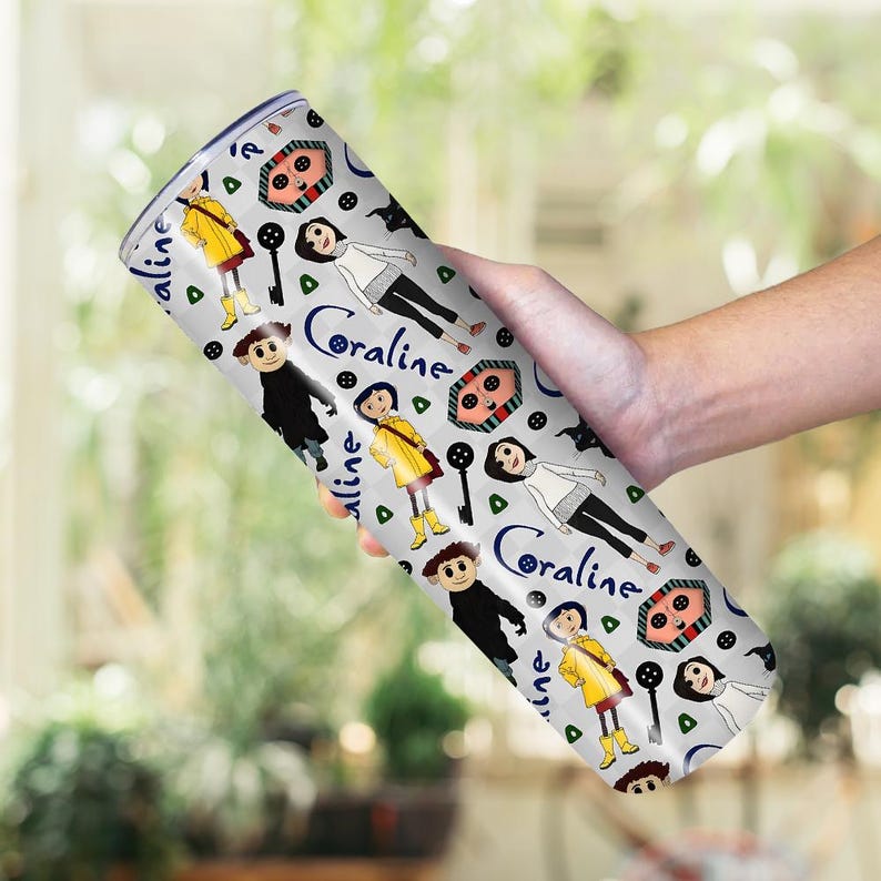 May include: A stainless steel tumbler featuring a repeating pattern of cartoon characters, keys, and buttons on a light gray background. The word "Coraline" is written in a blue script font. The tumbler is cylindrical and appears to be approximately 8 inches tall.