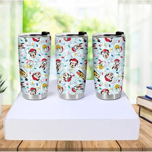 May include: Three stainless steel tumblers with a festive design. The tumblers feature a light blue background with images of Mickey Mouse, Donald Duck, and Goofy wearing Santa hats, along with Christmas lights.