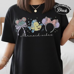May include: Black t-shirt with a colorful graphic featuring a cartoon fish, seashell ears, and the text "Mermaid vibes." The shirt is labeled "Comfort Colors Black."