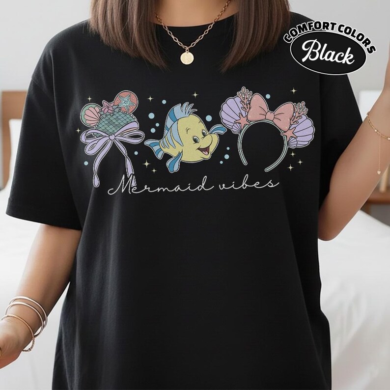 May include: Black t-shirt with a colorful graphic featuring a cartoon fish, seashell ears, and the text "Mermaid vibes." The shirt is labeled "Comfort Colors Black."