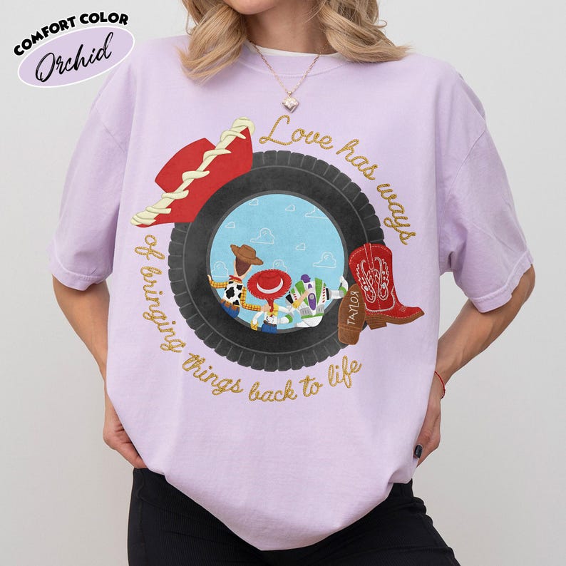 Może przedstawiać: Jasnofioletowy t-shirt z grafiką czarnej opony otaczającej scenę z niebieskim niebem i trzema postaciami z kresk&oacute;wek. Wz&oacute;r zawiera czerwony kapelusz kowbojski, czerwony but kowbojski oraz złoty napis Love has ways of bringing things back to life.