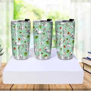 Puede incluir: Tres vasos de acero inoxidable con tapas negras. Cada vaso presenta un patr&oacute;n repetido de un mu&ntilde;eco de nieve, bastones de caramelo y regalos envueltos sobre un fondo verde claro. Los vasos son cil&iacute;ndricos y tienen una base plateada.