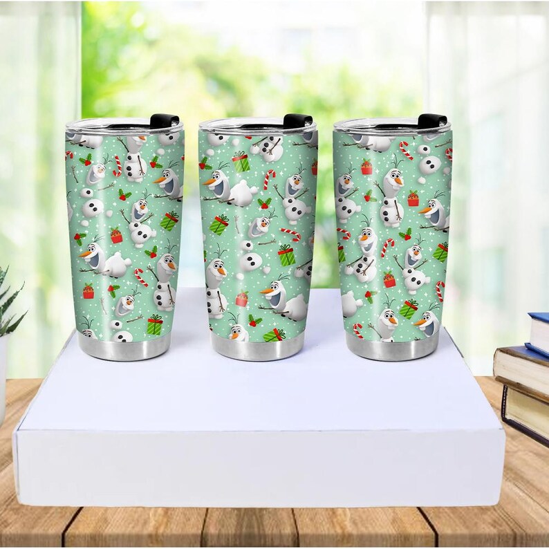 Puede incluir: Tres vasos de acero inoxidable con tapas negras. Cada vaso presenta un patr&oacute;n repetido de un mu&ntilde;eco de nieve, bastones de caramelo y regalos envueltos sobre un fondo verde claro. Los vasos son cil&iacute;ndricos y tienen una base plateada.