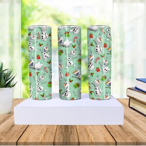 Puede incluir: Tres vasos cil&iacute;ndricos con un fondo verde claro est&aacute;n decorados con un patr&oacute;n de Olaf el mu&ntilde;eco de nieve, regalos envueltos y bastones de caramelo. Los vasos se exhiben sobre una plataforma blanca.