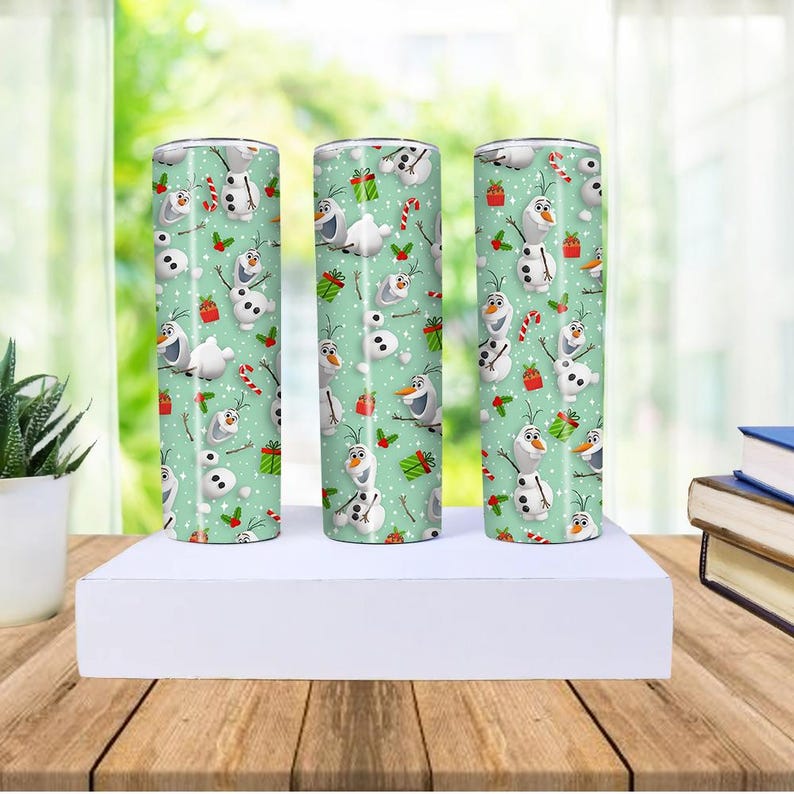 Puede incluir: Tres vasos cil&iacute;ndricos con un fondo verde claro est&aacute;n decorados con un patr&oacute;n de Olaf el mu&ntilde;eco de nieve, regalos envueltos y bastones de caramelo. Los vasos se exhiben sobre una plataforma blanca.