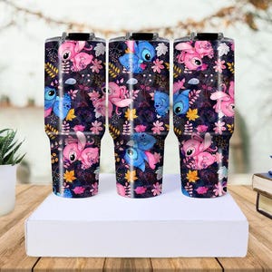 May include: Three stainless steel tumblers with a wraparound design featuring the character Stitch in pink and blue, set against a dark floral background. The tumblers are cylindrical with a black lid and handle.