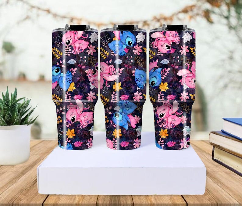 May include: Three stainless steel tumblers with a wraparound design featuring the character Stitch in pink and blue, set against a dark floral background. The tumblers are cylindrical with a black lid and handle.