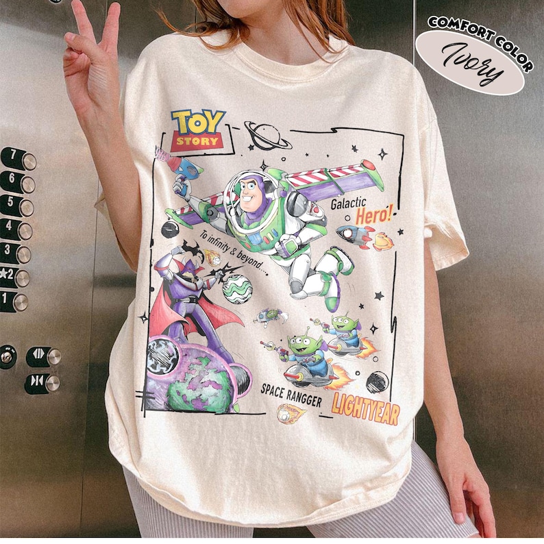 May include: An ivory t-shirt featuring a Toy Story graphic with Buzz Lightyear, Zurg, and green aliens. The print includes the text Toy Story, To infinity & beyond, Galactic Hero, Space Rangger, and Lightyear, surrounded by space elements.