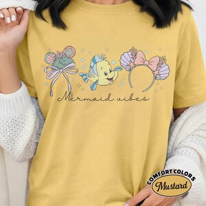 May include: Mustard-colored t-shirt with a graphic featuring a cartoon fish, a seashell headband with a pink bow, and a decorative bow. The text "Mermaid vibes" is written in cursive. The shirt is labeled "Comfort Colors".