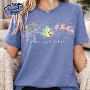 May include: Blue t-shirt with the text "Mermaid vibes" and illustrations of mermaid-themed headbands and a yellow fish. The shirt has a "Comfort Colors" and "Washed Denim" logo in the upper left corner.
