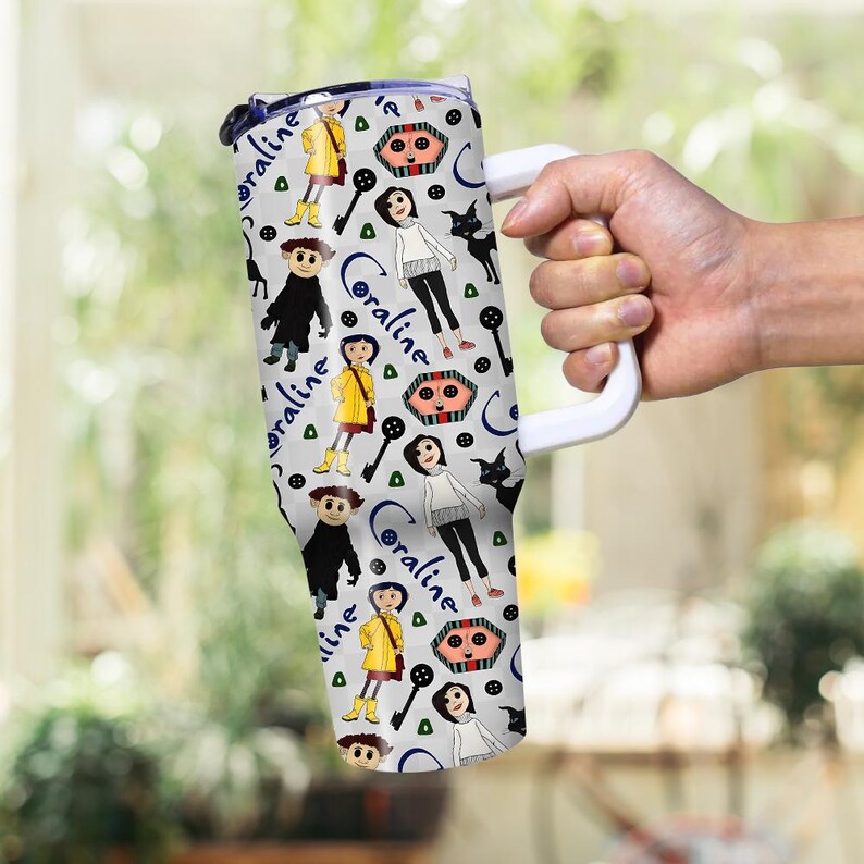 May include: A white tumbler with a wraparound pattern featuring characters and elements from the movie "Coraline." The design includes illustrations of Coraline, the Other Mother, black cats, keys, and the word "Coraline" in a stylized font. The tumbler has a white handle.