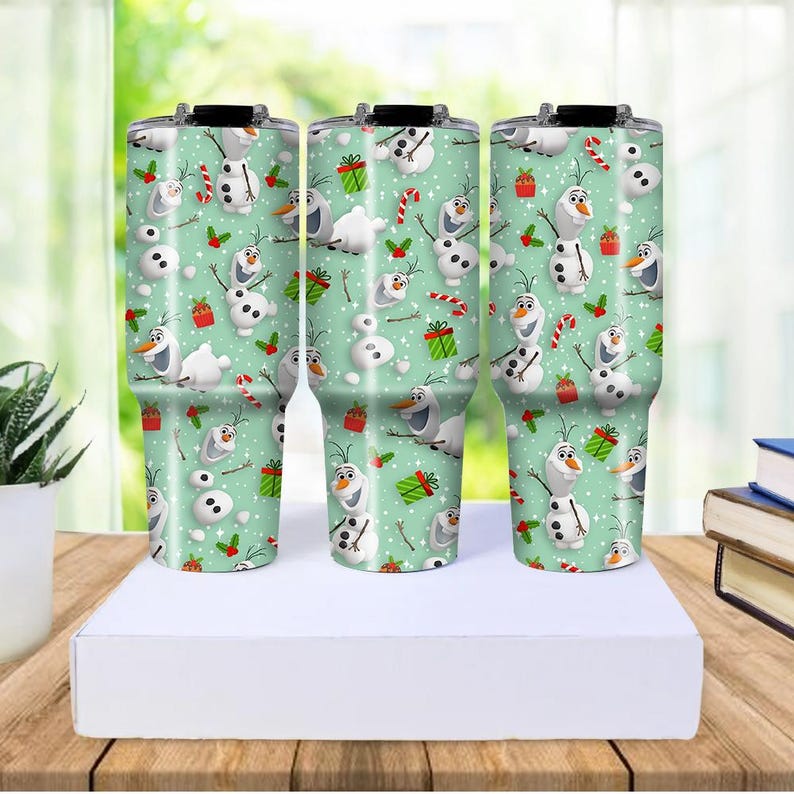 Puede incluir: Tres vasos aislados con un fondo verde menta y un patr&oacute;n repetido de Olaf el mu&ntilde;eco de nieve, bastones de caramelo y regalos envueltos. Los vasos son de acero inoxidable con tapas negras.