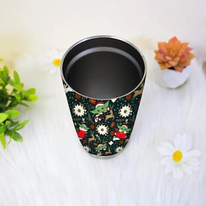 Pode incluir: Um copo de a&ccedil;o inoxid&aacute;vel com fundo verde escuro e um padr&atilde;o repetido de um personagem de desenho animado com chap&eacute;u de Papai Noel, renas e flocos de neve. O copo cil&iacute;ndrico tem uma borda prateada.