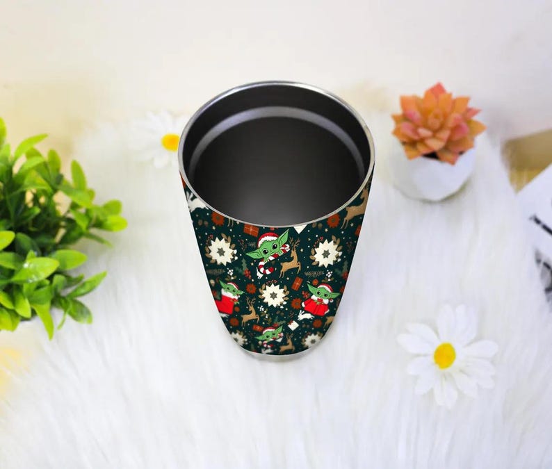 Pode incluir: Um copo de a&ccedil;o inoxid&aacute;vel com fundo verde escuro e um padr&atilde;o repetido de um personagem de desenho animado com chap&eacute;u de Papai Noel, renas e flocos de neve. O copo cil&iacute;ndrico tem uma borda prateada.
