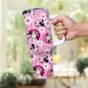 以下が含まれることがあります： ミニーマウスの顔、白いデイジー、ピンクの花柄が描かれたピンク色のタンブラー。タンブラーには白いハンドルがあり、側面には「ミニー」の文字がプリントされています。タンブラーは人が持っています。