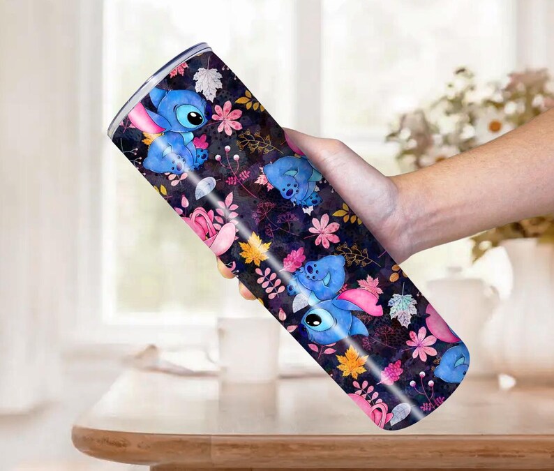 May include: A stainless steel tumbler featuring a repeating pattern of the character Stitch from Lilo & Stitch. The design includes blue Stitch figures, pink and yellow flowers, and leaves on a dark background.