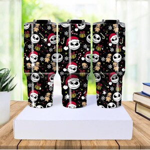 May include: Three black insulated tumblers with a festive pattern featuring Jack Skellington in a Santa hat, gifts, snowflakes, and candy canes. The tumblers are on a white surface, with a wooden table and greenery in the background.