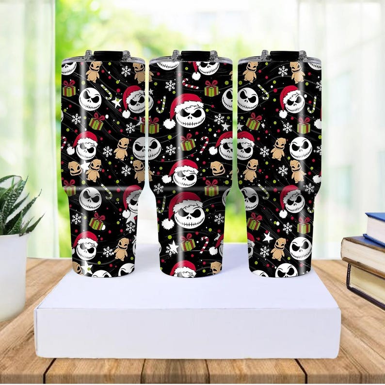 May include: Three black insulated tumblers with a festive pattern featuring Jack Skellington in a Santa hat, gifts, snowflakes, and candy canes. The tumblers are on a white surface, with a wooden table and greenery in the background.