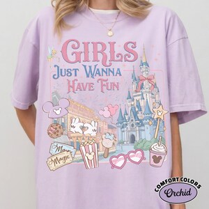 May include: A light purple t-shirt with the words "GIRLS JUST WANNA HAVE FUN" in pink and blue. The shirt features a graphic of a castle, Mickey Mouse, and other theme park icons. The shirt is a Comfort Colors Orchid.