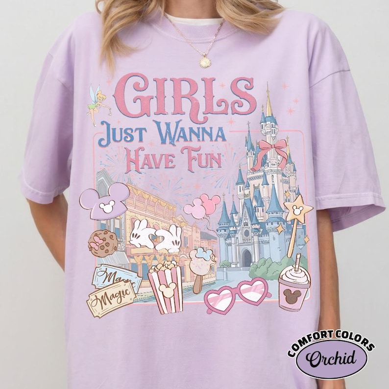 May include: A light purple t-shirt with the words "GIRLS JUST WANNA HAVE FUN" in pink and blue. The shirt features a graphic of a castle, Mickey Mouse, and other theme park icons. The shirt is a Comfort Colors Orchid.