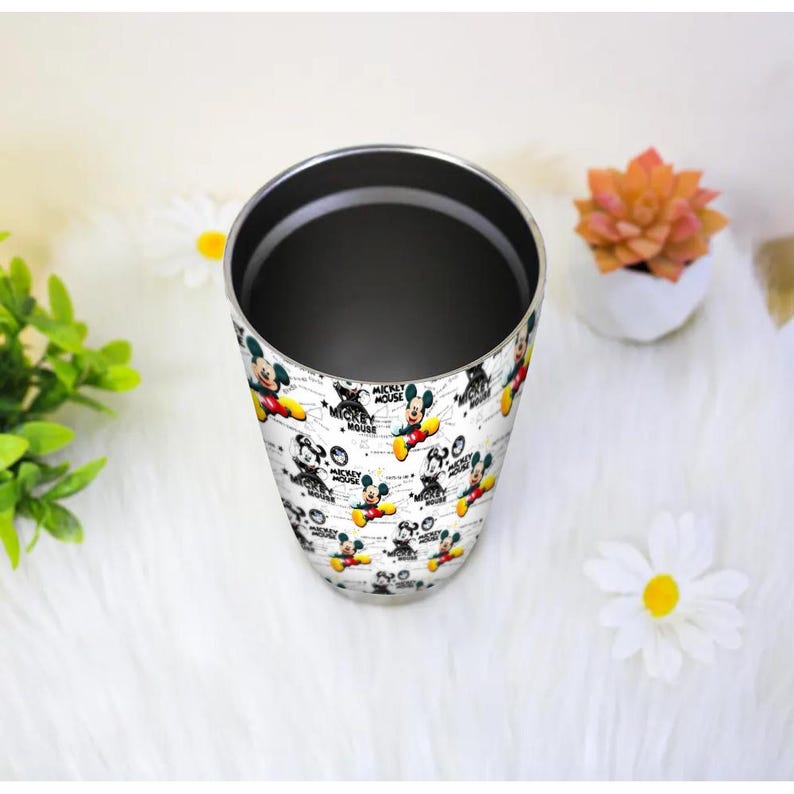 May include: A stainless steel tumbler featuring a repeating pattern of Mickey Mouse in various poses. The design includes the text "Mickey Mouse" in a playful font, set against a white background. The tumbler has a silver interior and a smooth, glossy finish.