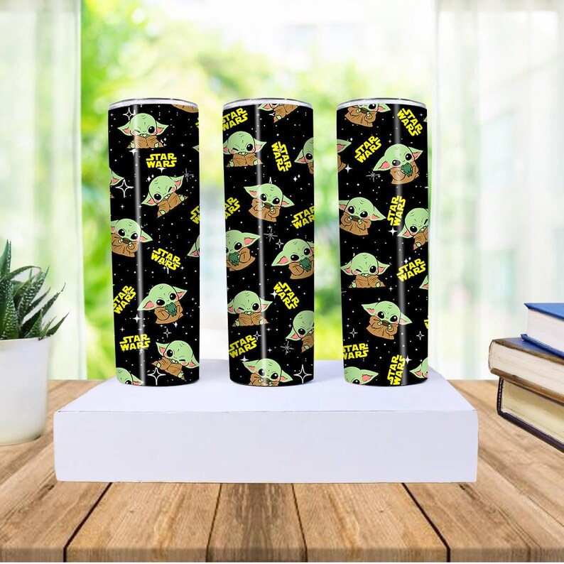 May include: Three stainless steel tumblers with a black background, featuring a repeating pattern of a green alien character and the words "STAR WARS" in yellow. The tumblers are cylindrical and sit on a white platform.