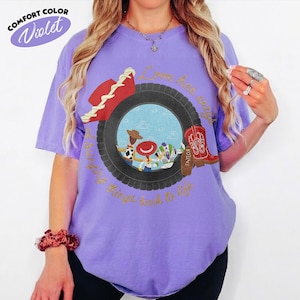 Może przedstawiać: Fioletowy t-shirt z kr&oacute;tkim rękawem z grafiką czarnej opony otaczającej scenę z trzema postaciami z kresk&oacute;wek. Napis "Love has ways of bringing things back to life" jest wydrukowany złotą czcionką. Widoczny jest r&oacute;wnież czerwony kapelusz i czerwony kowbojski but.