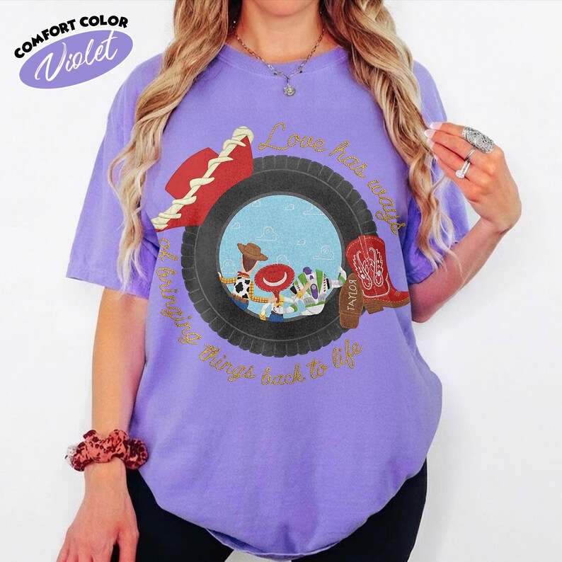 Może przedstawiać: Fioletowy t-shirt z kr&oacute;tkim rękawem z grafiką czarnej opony otaczającej scenę z trzema postaciami z kresk&oacute;wek. Napis "Love has ways of bringing things back to life" jest wydrukowany złotą czcionką. Widoczny jest r&oacute;wnież czerwony kapelusz i czerwony kowbojski but.