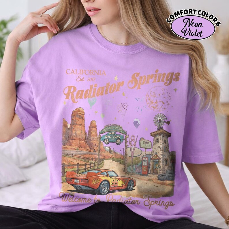 Puede incluir: Una camiseta de color lila con un gr&aacute;fico de Radiator Springs, California. El dise&ntilde;o incluye un coche de carreras rojo, un paisaje des&eacute;rtico y el texto "Radiator Springs" y "Welcome to Radiator Springs". La camiseta tambi&eacute;n tiene el texto "COMFORT COLORS Neon Violet".