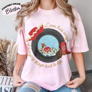Może przedstawiać: Jasnor&oacute;żowy t-shirt z kr&oacute;tkim rękawem z grafiką przedstawiającą czarną oponę otaczającą scenę z trzema postaciami z kresk&oacute;wek. Wok&oacute;ł opony nadrukowany jest złoty napis "Love has ways of bringing things back to life" oraz czerwony kapelusz i but.