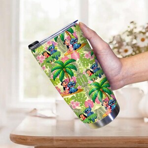May include: A stainless steel tumbler featuring a tropical design with cartoon characters, palm trees, and pink flowers. The tumbler has a black lid and is held by a person.
