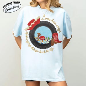Może przedstawiać: Jasnoniebieski t-shirt z grafiką czarnej opony otaczającej postacie z kresk&oacute;wek. Wz&oacute;r zawiera czerwony kowbojski kapelusz, czerwony but oraz napis "Love has ways of bringing things back to life" wykonany złotą kursywą.