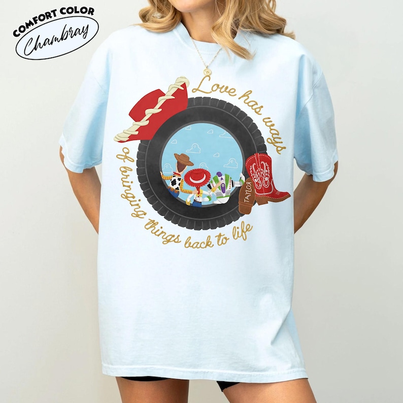Może przedstawiać: Jasnoniebieski t-shirt z grafiką czarnej opony otaczającej postacie z kresk&oacute;wek. Wz&oacute;r zawiera czerwony kowbojski kapelusz, czerwony but oraz napis "Love has ways of bringing things back to life" wykonany złotą kursywą.