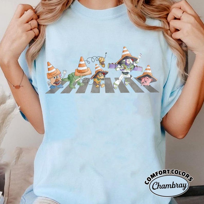 K&ouml;nnte beinhalten: Hellblaues T-Shirt mit einer verspielten Illustration von Zeichentrickfiguren, darunter Mr. Potato Head und Buzz Lightyear, die einen Zebrastreifen &uuml;berqueren, wobei jede Figur einen Verkehrskegelhut tr&auml;gt. Das Shirt hat das Comfort Colors Chambray Logo.