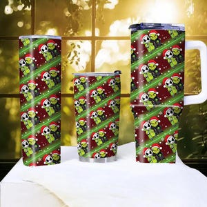 May include: A set of three insulated tumblers with a festive Christmas design. The tumblers feature a repeating pattern of green characters wearing Santa hats, set against a red background with green diagonal stripes and snowflakes. One tumbler has a handle.