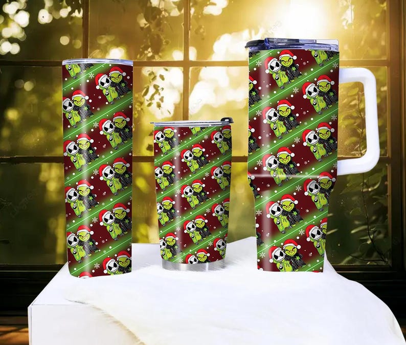 May include: A set of three insulated tumblers with a festive Christmas design. The tumblers feature a repeating pattern of green characters wearing Santa hats, set against a red background with green diagonal stripes and snowflakes. One tumbler has a handle.