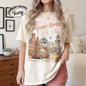 Puede incluir: Camiseta marfil Comfort Colors con un gr&aacute;fico de Radiator Springs, California. El dise&ntilde;o incluye un coche de carreras rojo, un paisaje des&eacute;rtico y el texto "Welcome to Radiator Springs". La camiseta tiene un aspecto vintage.