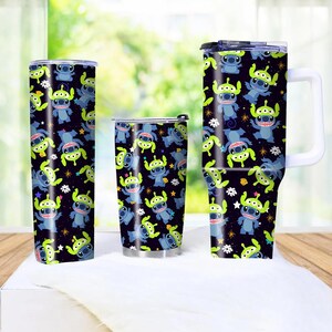 Peut inclure: Ensemble de trois gobelets isothermes avec un motif r&eacute;p&eacute;t&eacute; d'extraterrestres de dessins anim&eacute;s bleus et verts sur fond noir. Les gobelets sont de tailles diff&eacute;rentes, dont un grand gobelet de voyage, un gobelet standard et une tasse avec une poign&eacute;e.