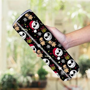 May include: A black, cylindrical tumbler with a festive design. The pattern includes Jack Skellington in a Santa hat, Oogie Boogie, snowflakes, and wrapped gifts. The tumbler is likely made of stainless steel.