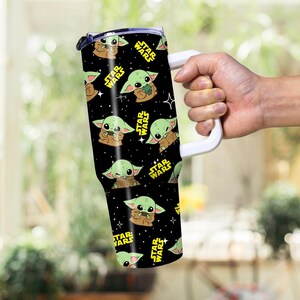 May include: A black tumbler with a repeating pattern of Grogu from Star Wars, and the Star Wars logo in yellow. The tumbler has a white handle and a black lid. The background is blurred.